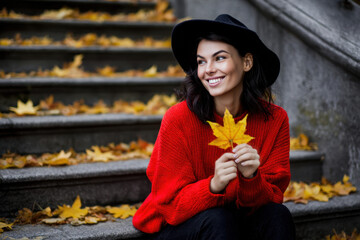 A woman with dark hair sits on stone steps covered in yellow leaves, holding a maple leaf and smiling brightly as the sun shines