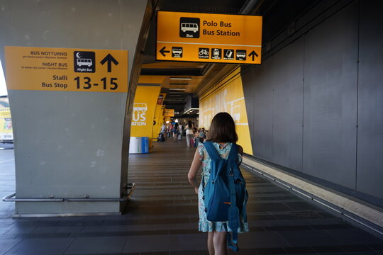 At Fiumicino Airport a young girl with a blue backpack walks toward the brightly signed Polo Bus Station area, heading to bus stops for city and night routes - Blurred people in the background.