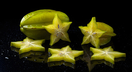 Still life of star fruit: a whole carambola and several star-shaped slices arranged on a reflective black surface.
