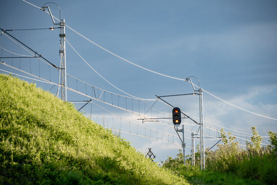 Modern railway traction system with red signal light, set against vibrant green grass and a dramatic cloudy sky. A clean blend of technology and nature captured in one frame