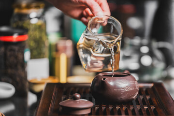 Hand pouring hot water from a glass pitcher into a brown teapot on a wooden tray. Cozy tea setup with jars in background. Traditional tea ceremony atmosphere.