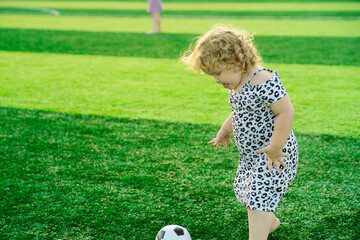 A young girl with curly hair happily kicks a soccer ball on a lush green field under bright...