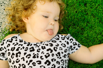 A young girl with curly hair is lying on green grass playfully sticking out her tongue She wears a...