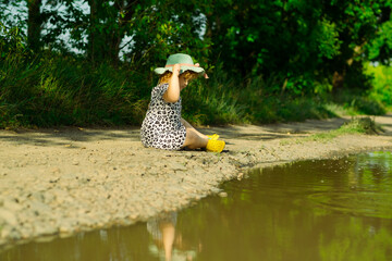 A young child wearing a hat and a patterned dress joyfully throws pebbles into a puddle along a...