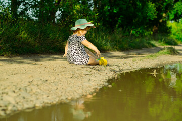 A young child wearing a hat and a patterned dress joyfully throws pebbles into a puddle along a...