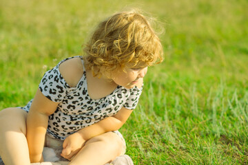 A young child with curly hair sits on green grass wearing a light outfit The child appears...