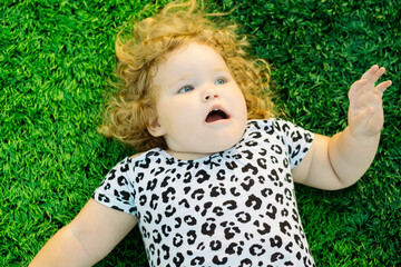 A young child lies on vibrant green grass with curly hair framing a surprised expression The sun...