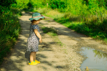A young child in a playful dress and hat tosses pebbles into a puddle watching the ripples form The...