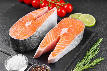 Two fresh raw salmon or trout steaks on black stone surface with food ingredients. Tomatoes, lime,...