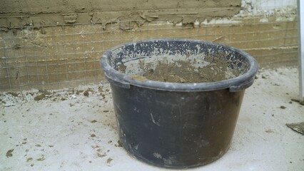 Black bucket containing mortar mix placed on a construction site floor, with a partially plastered wall and wire mesh in the background