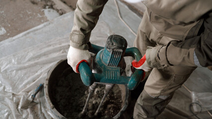 Construction worker wearing protective gloves mixing cement with electric mixer in bucket at construction site