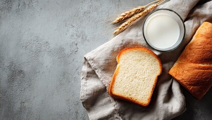 Bread loaf, slice, milk glass, and wheat stalks on a linen cloth and grey textured surface