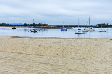 Empty sandy beach at Mariners Basin full of moored boats and yachts on a cloudy summer morning, San...