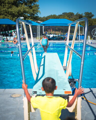 Mid-jump girl sails toward the water as a motion blurry boy in green watches from behind. The scene radiates excitement and peer connection beneath sunny skies and colorful shade canopies, Texas