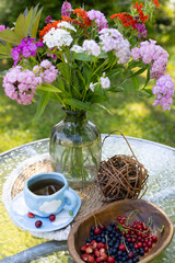 On the glass table there is a vase with bright flowers, a cup of coffee and a vase with assorted berries. A bright and cozy environment in nature.