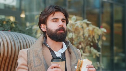 Indian young businessman tourist enjoy lunch break eating tasty sandwich meal and drinking hot beverage coffee or tea. Arabian bearded man guy relaxing sitting on a bench on downtown city street - Powered by Adobe