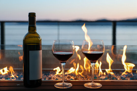 A nice bottle of red wine with two glasses next to a comfortable file with orange flames. A beautiful lake or ocean in the background with the breathtaking view from the vacation home porch. 