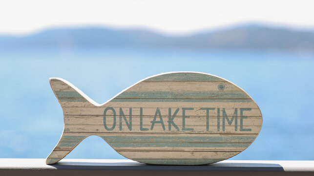 A small fish shaped wooden sign that says On Lake Time outside on a hot summer day near the water. 