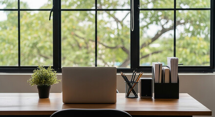 Fototapeta premium Bright Workspace Featuring Laptop, Potted Plant, and Office Supplies on a Wooden Desk in Front of a Large Window
