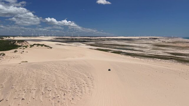 A buggy is driving across the Canoa Quebrada dunes, and wind turbines can be seen in the distance.