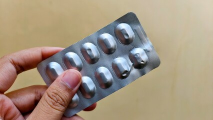 Hand holding a blister pack of medication, some pills missing, against a light brown background,...