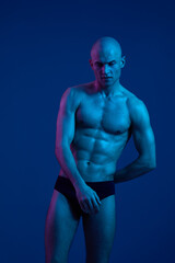 Athletic man posing in blue lighting, showcasing strong physique and confident expression. The...