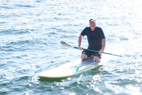Happy middle aged man resting on his knees on a paddle board outside on a lake. He is holding a paddle over the water and working to maintain balance. 