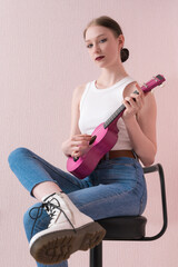 Woman musician strumming ukulele and looking at camera. Woman skillfully plucking stringed musical instrument, sitting on bar stool with legs crossed at knees. Woman in white vest, jeans and boots