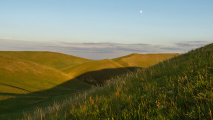 Rolling green hills under a clear blue sky with soft shadows cast by the landscape, showcasing the beauty of nature and tranquility in a serene outdoor environment