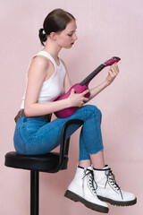 Woman holding ukulele and tuning strings of musical instrument with concentration. Profile view of musician sitting on bar stool. Performer is woman exuding confidence, wearing tank top, jeans, boots