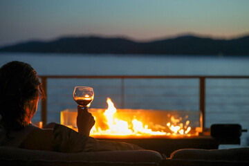 A woman sitting in an elegant setting at night outside drinking a glass of red wine with a warm fire overlooking a lake view. 