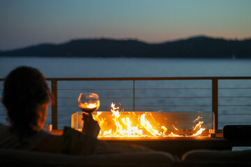 A woman sitting in an elegant setting at night outside drinking a glass of red wine with a warm fire overlooking a lake view. 