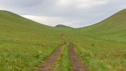 Obraz premium Winding dirt path through lush green hills under a cloudy sky, inviting exploration and adventure in a serene natural landscape with rolling terrain and vibrant grass