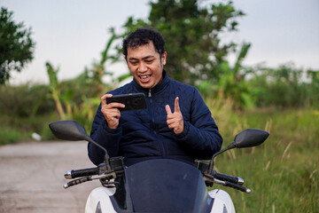 Happy young Asian man on a scooter enjoying entertainment on his smartphone outdoors in nature.
