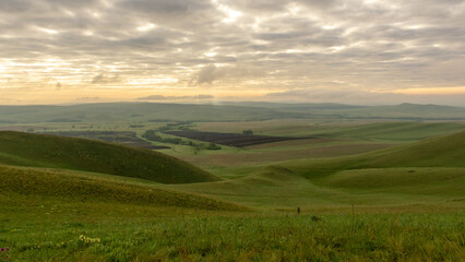 Expansive green hills under a cloudy sky at dawn, showcasing rolling landscapes and distant fields, creating a serene and tranquil atmosphere in nature's beauty