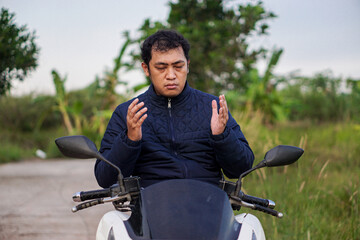 Anxious middle-aged Asian man sitting on his scooter on a country road, making a gesture of prayer or pleading.