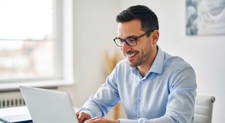 Smiling man in glasses wearing a blue shirt working on a laptop in an office environment