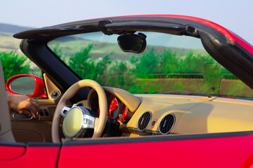 Horizontal photo. Interior of red, scarlet, crimson sport convertible car on nature. View of mountain, hill, bush. Steering wheel, windshield, rearview mirror, panel. Rent, transport, travel, tourism