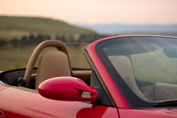 Horizontal street photo. Fragment of red, scarlet, crimson sport convertible car against background of mountain, hill. Windshield, rearview mirror, seat. Concept of transport, travel, tourism, rent