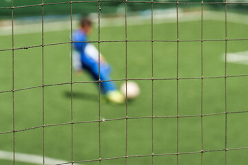 A soccer goal net in sharp focus, with a blurred background showing a player in a blue uniform about to kick the ball on a green artificial turf field