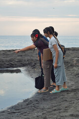 Standing on the beach, the girls look toward the brackish water while one girl gestures, possibly...