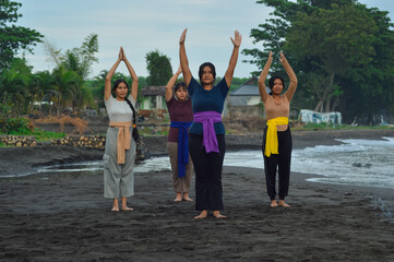 The teens perform a standing yoga pose with palms joined above their heads, facing the sea,...