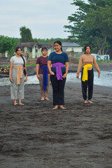 One girl gestures a cue to start yoga, standing apart with others near the river mouth on a quiet beach.
