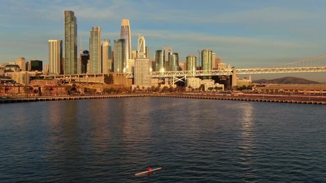 A lone kayaker paddles on the bay in San Francisco, California, USA, with the city skyline and Bay Bridge in the background during a sunny day.