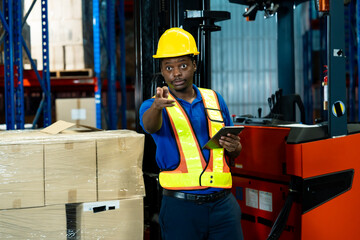 African male warehouse worker in safety vest and helmet smiling while pointing and holding digital...