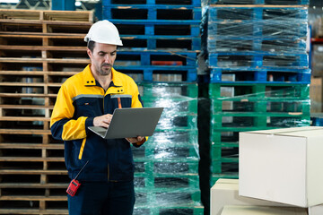 Male logistics staff in yellow blue uniform using laptop computer to update warehouse stock beside...