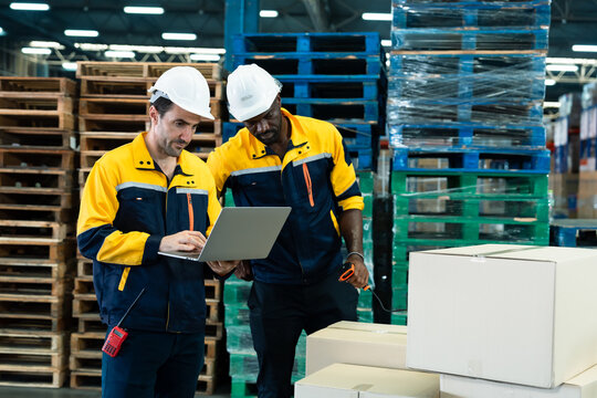 Two male warehouse workers analyzing delivery data together using laptop and scanner inside packaging section of logistics center showing multicultural teamwork and digital inventory management