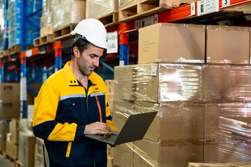 Caucasian male logistics worker monitoring warehouse stock using laptop wearing helmet and...