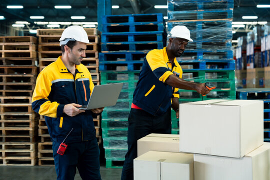 African male warehouse worker scanning cardboard box while caucasian colleague checks shipping record on laptop showing teamwork in inventory management inside pallet-loaded logistics facility