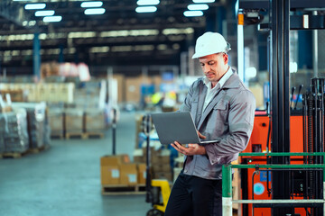 Caucasian male engineer in gray jacket and white helmet using laptop near forklift managing...